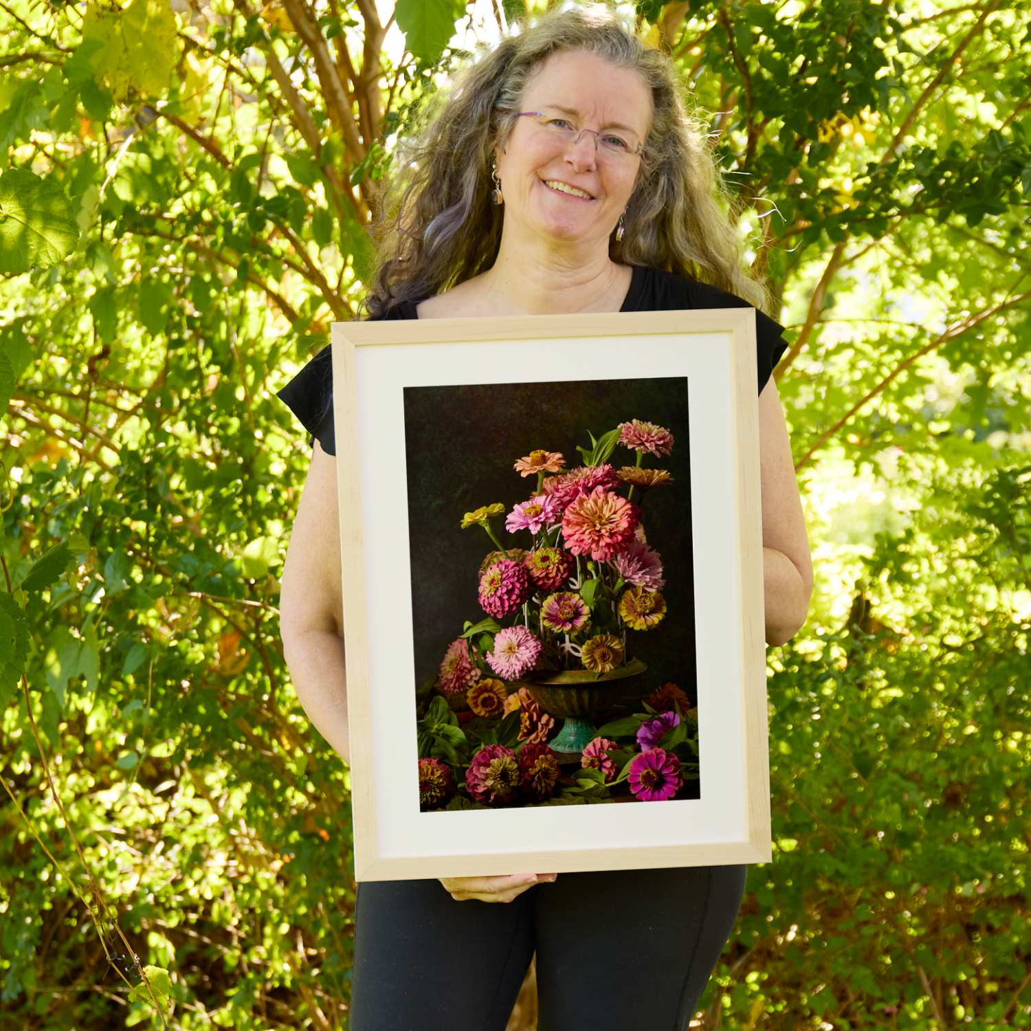 Christina Peters holding Petals In Flight Zinnia Fine Art Print, a dramatic still life of pink, coral and magenta zinnias in a footed teal bowl, with blooms spilling below against a deep black backdrop, framed in natural wood with a white mat. She is standing outdoors in front of sunlit green trees and leafy shrubs.