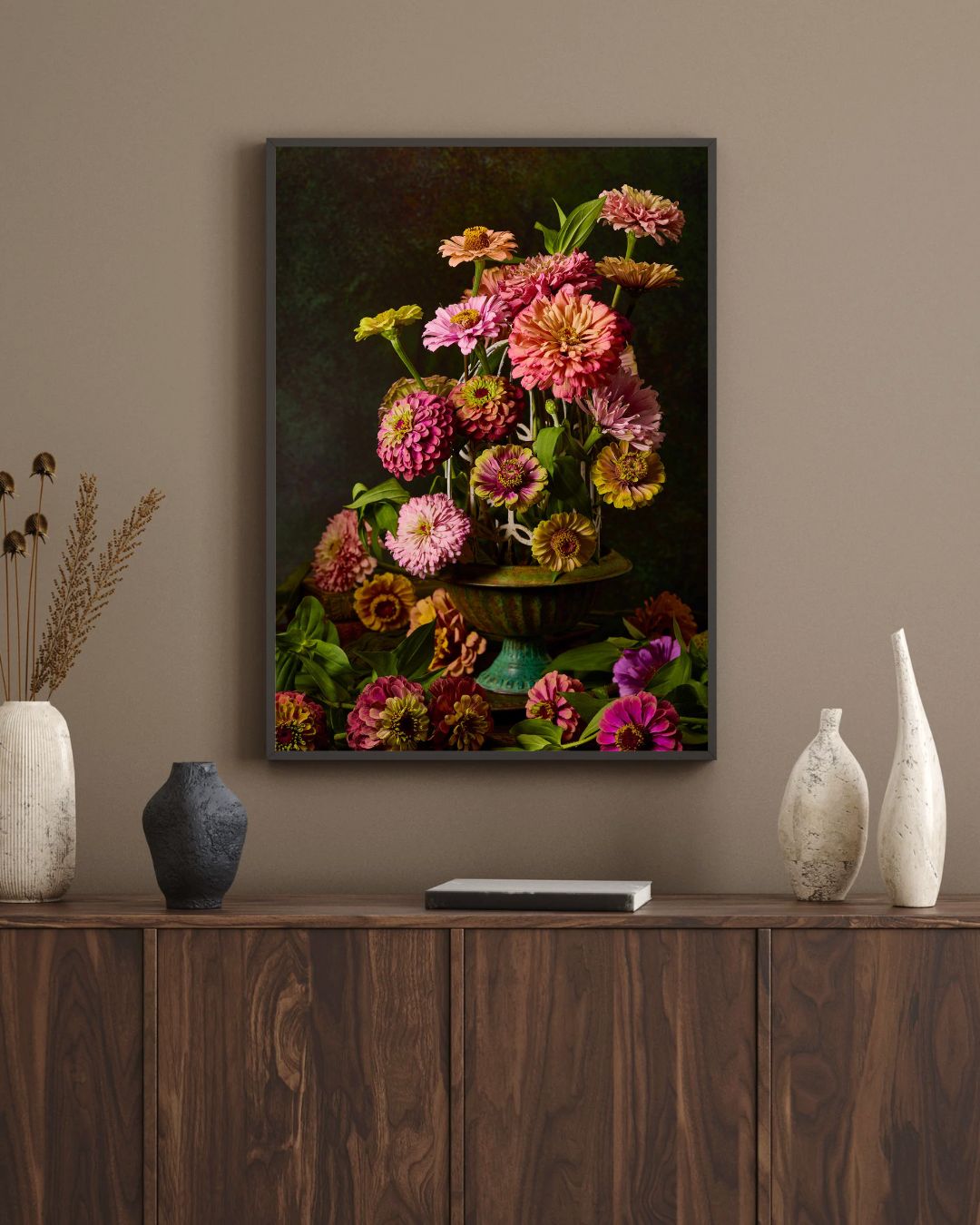 The framed canvas Petals In Flight adds a vibrant touch to the wall. Below, a wooden sideboard showcases three decorative vases in white, black, and gray. A textured vase with dried grass stands on the left against a soft beige background.