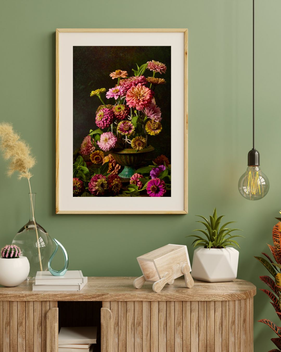 A stylish room with green walls showcases the Petals In Flight art print of zinnias, a hanging light bulb on the right, and a wooden table adorned with decor, books, and plants. On the left, a vase holds dried grasses.