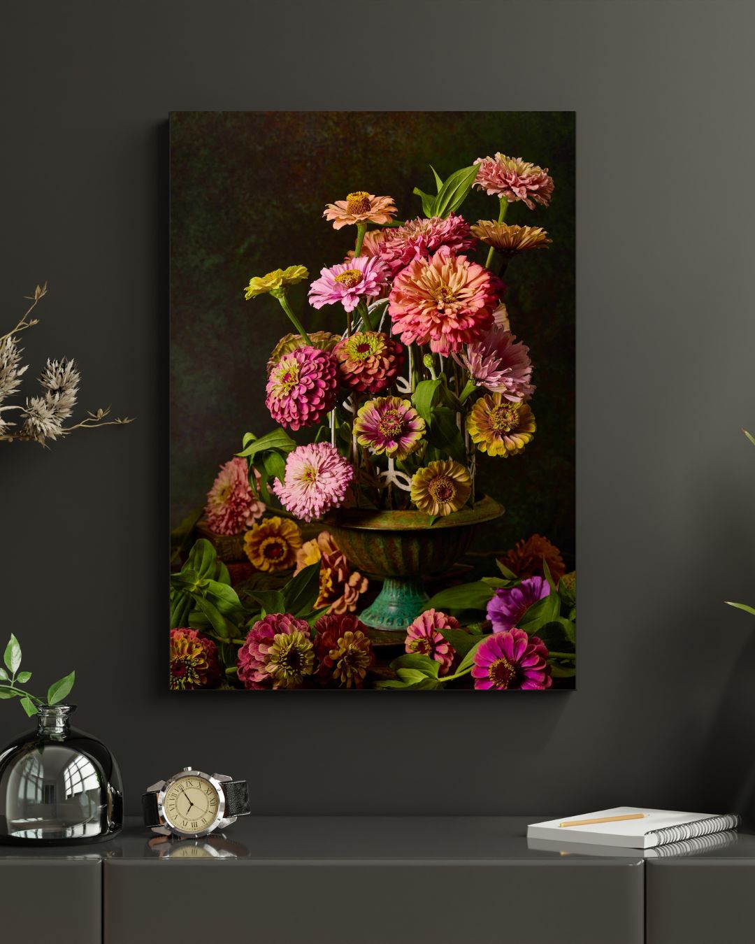 The wall-mounted canvas art Petals In Flight features a vibrant still-life of colorful zinnias in a green vase, accompanied by a small plant, clock, and book on a table against a dark wall.