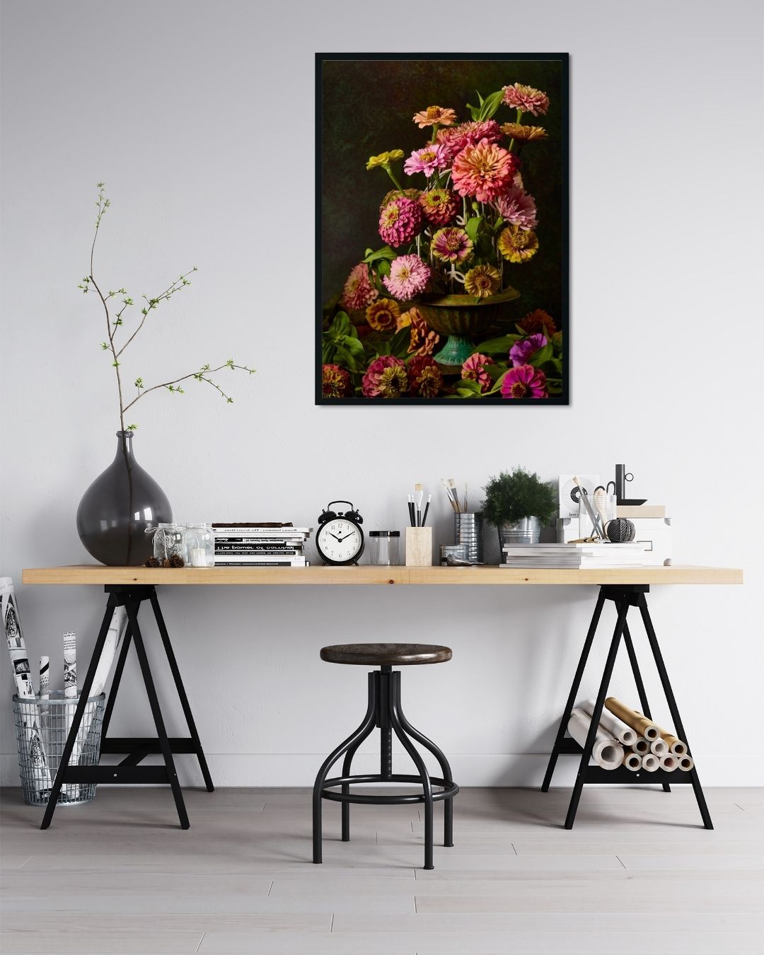 In a minimalist workspace, a wooden desk with a black metal base holds an alarm clock, books, and potted plants. A stool stands before it, while the vibrant Petals In Flight canvas wall art depicting zinnias hangs on the white wall above, adding color.