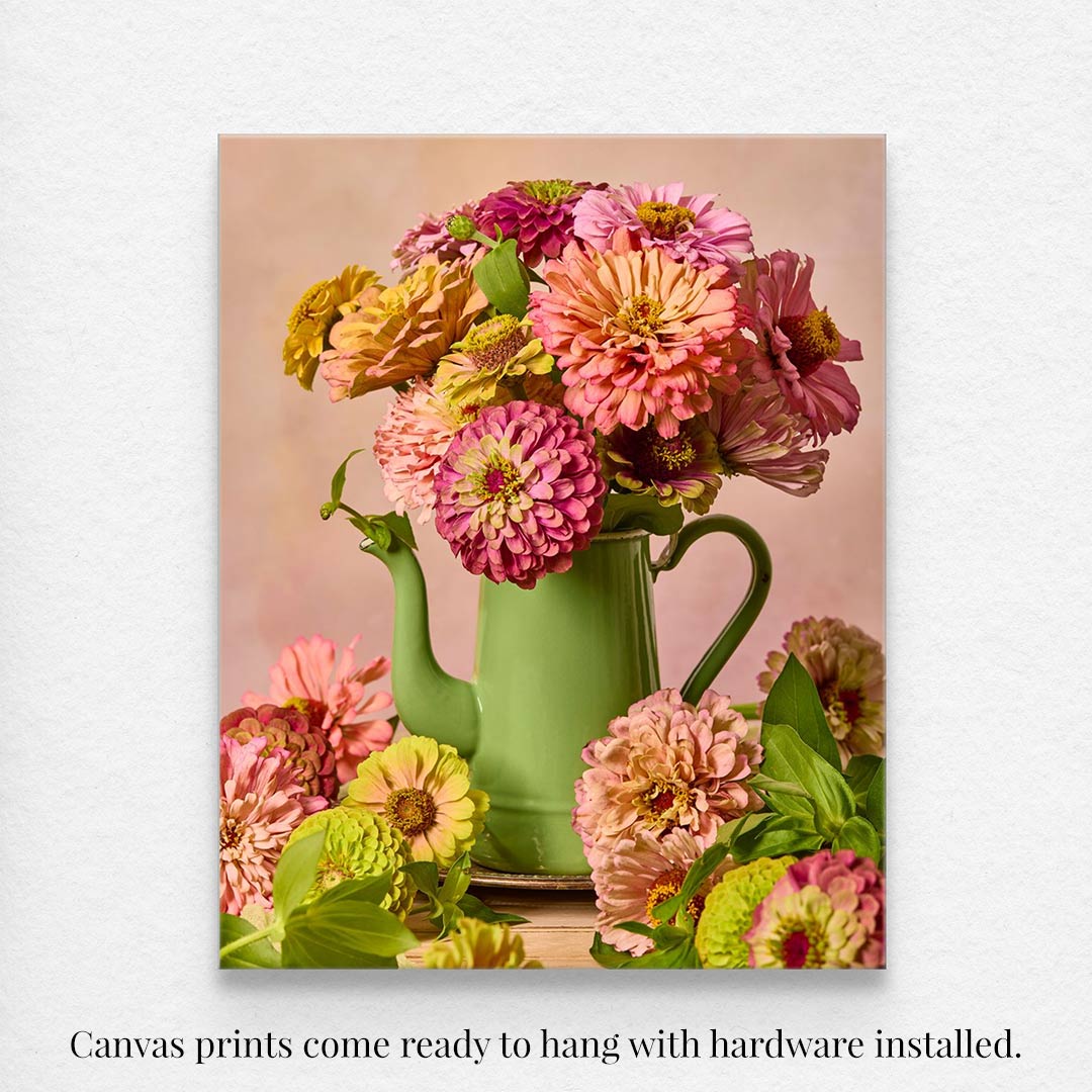 The canvas wall art, Steeping Beauty, showcases a vintage green teapot overflowing with pink and orange flowers amidst more blossoms against a soft background. It comes ready to hang with hardware installed.