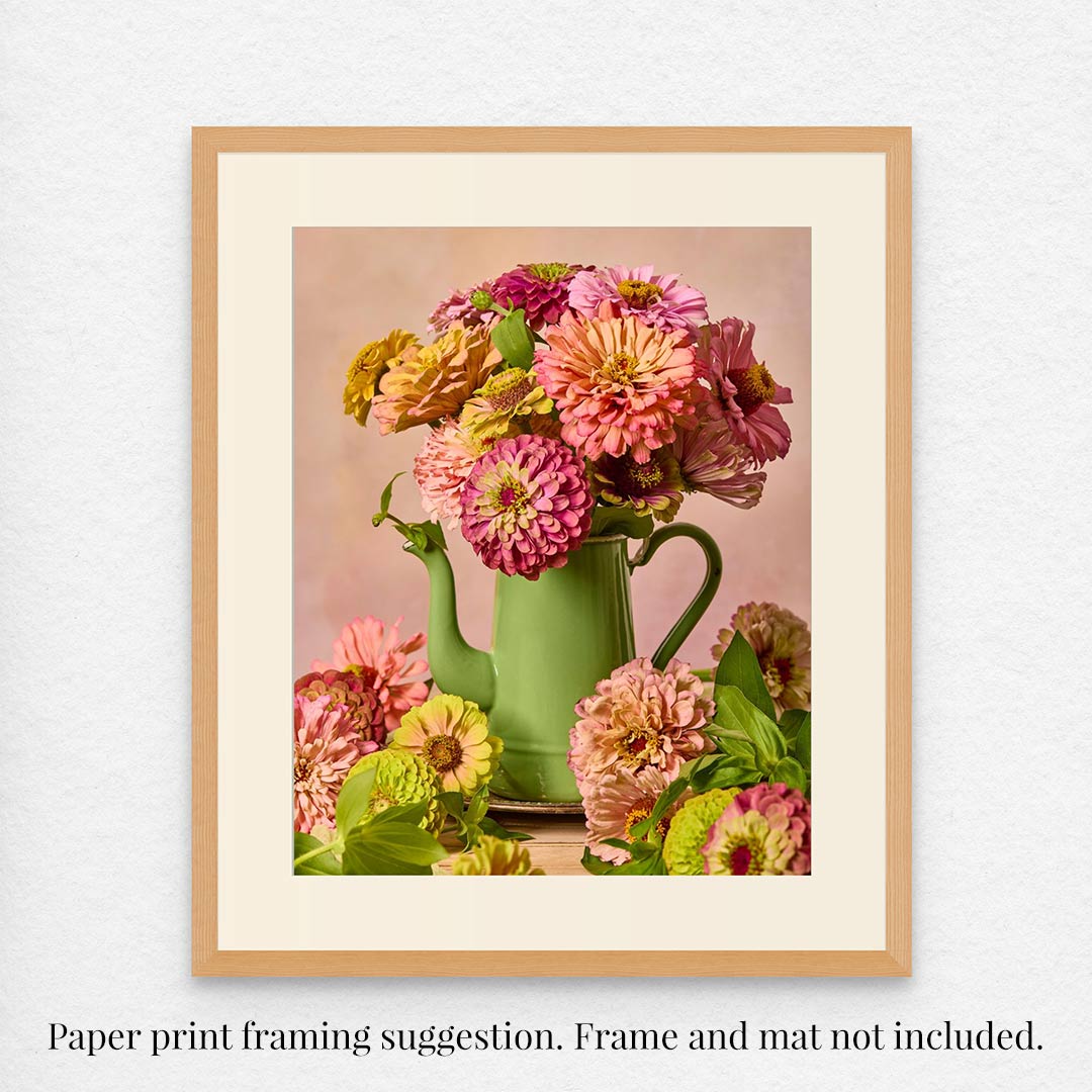 The Steeping Beauty print showcases a vintage green teapot vase adorned with pink and orange flowers, with scattered blossoms. It features a light wood frame and white mat. Note: Frame and mat are not included; this is only a framing suggestion.