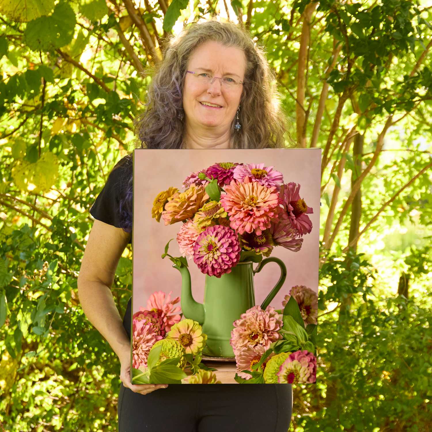 Christina Peters holding Steeping Beauty Zinnia Wall Art Canvas, a still life of coral, rose and pale green zinnias in a sage green enamel teapot, with blooms gathered at its base against a soft pink backdrop. She is standing outdoors in front of sunlit green trees and leafy shrubs.