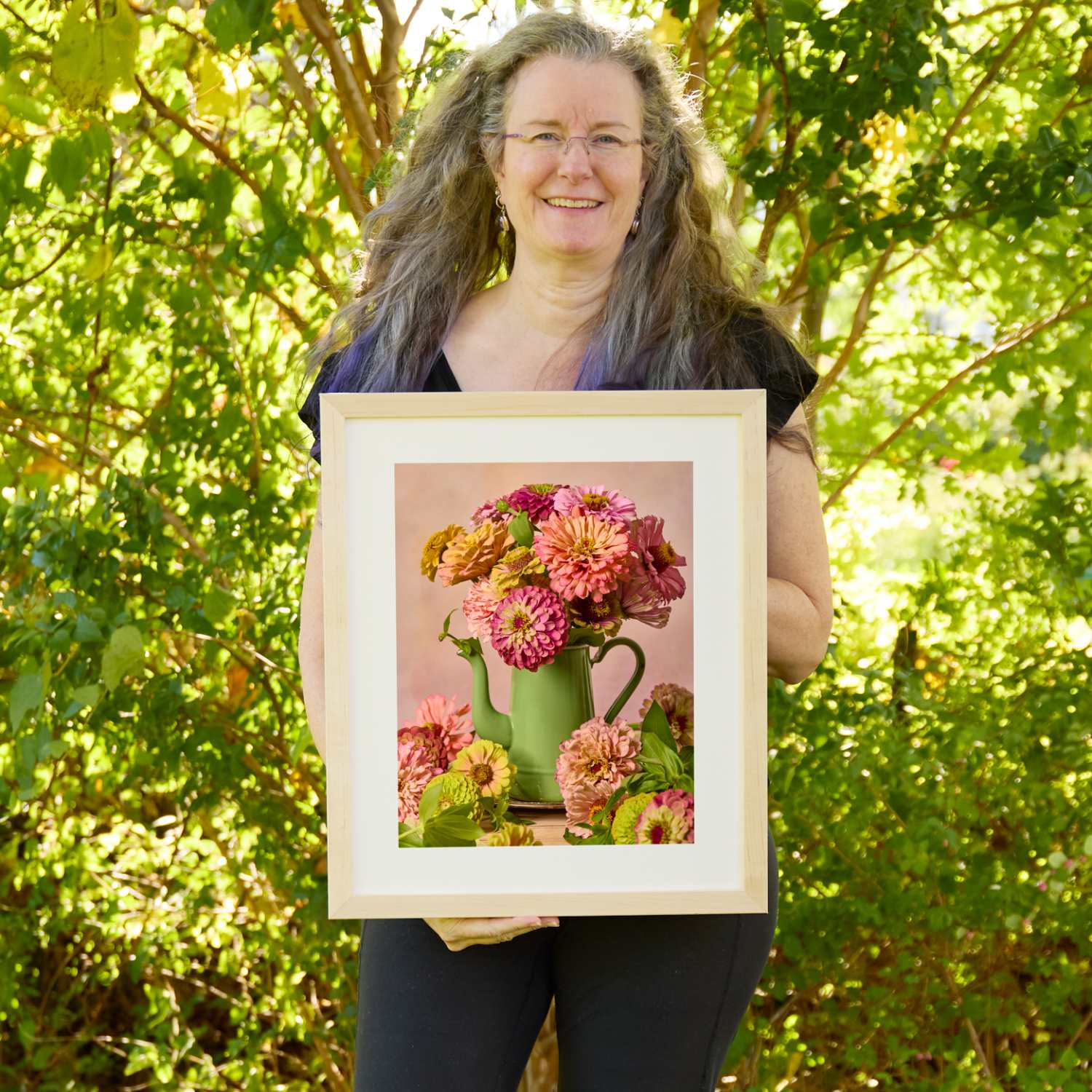 Christina Peters holding Steeping Beauty Zinnia Wall Art Print, a still life of coral, rose and pale green zinnias in a sage green enamel teapot, with blooms gathered at its base against a soft pink backdrop, framed in natural wood with a white mat. She is standing outdoors in front of sunlit green trees and leafy shrubs.