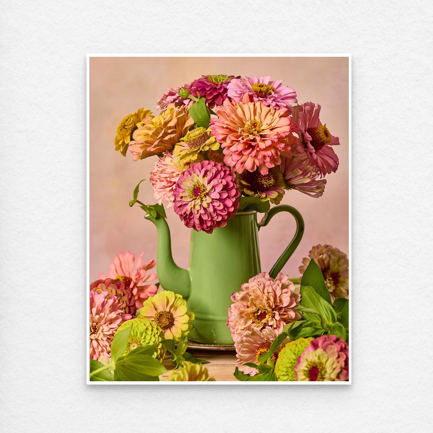 The Steeping Beauty bouquet of vibrant pink, yellow, and orange zinnias in a vintage green teapot creates an artistic scene perfect for canvas wall art. Scattered blooms lie around the teapot on a wooden surface, all set against a soft, neutral background.