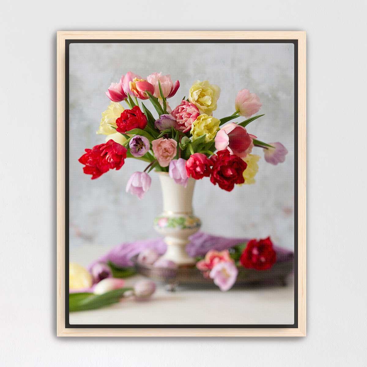 A white vase filled with a vibrant bouquet of red, pink, yellow, and white flowers sits on a table, surrounded by additional flowers and leaves.