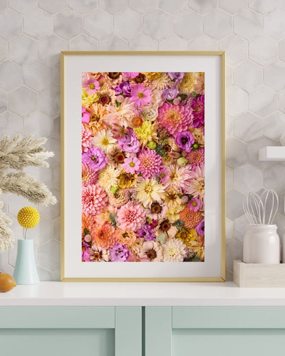 The framed paper print Christinas Wedding, featuring colorful flowers like pink, yellow, and purple blossoms, leans on a hexagonal tile-patterned wall. Below it, a white countertop displays decorative vases and a glass, with pale green cabinets below.