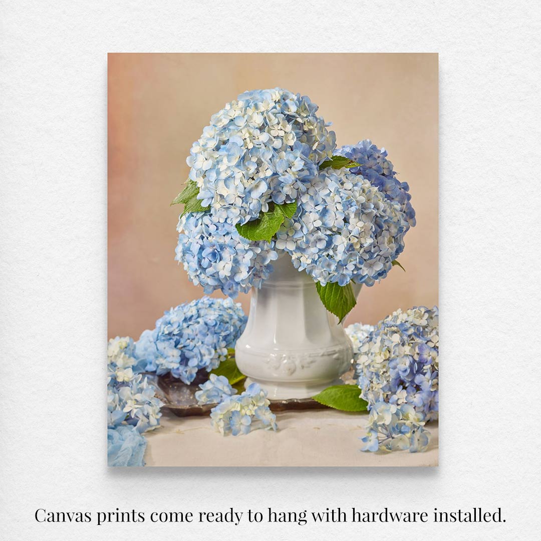 The Dreaming of Something Blue canvas art captures blue hydrangeas in a white vase with a soft background, with extra flower beside the vase. The artwork comes ready to hang with hardware installed.