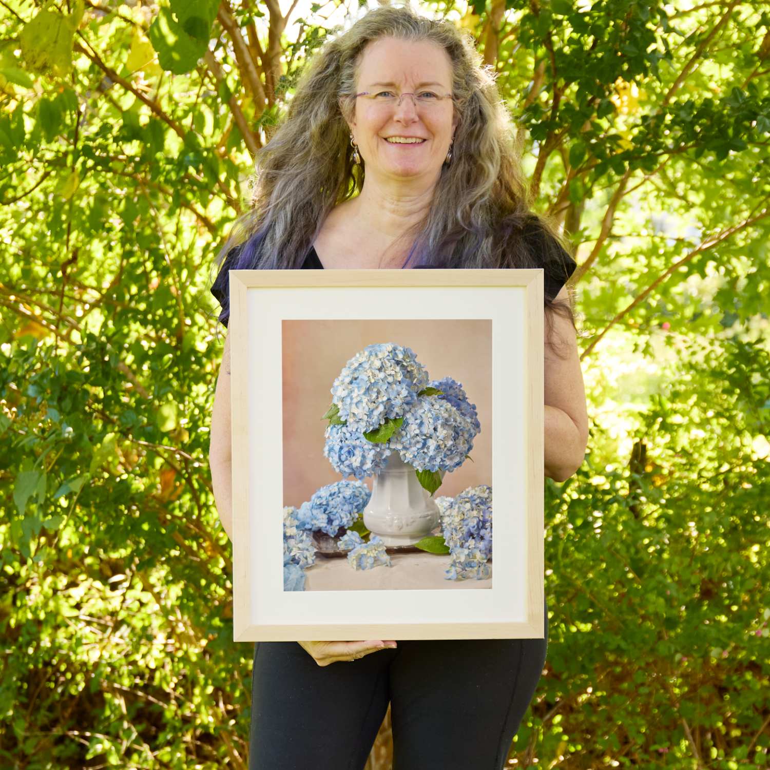 Christina Peters holding Dreaming of Something Blue Hydrangea Wall Art Print, a still life of pale blue hydrangeas in a white ironstone urn, with loose blooms around its base against a soft blush backdrop, framed in natural wood with a white mat. She is standing outdoors in front of sunlit green trees and leafy shrubs.