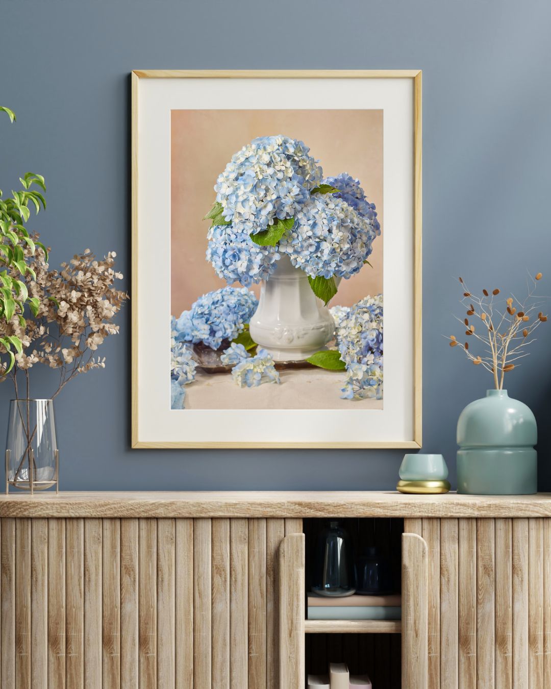 The Dreaming of Something Blue framed print, featuring blue hydrangeas in a white vase, hangs on a blue wall. Below it, a wooden cabinet displays decorative items such as vases and dried plants. Mat and frame not included.