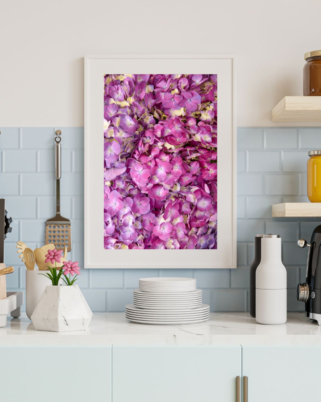 A kitchen counter with a blue tile backsplash. On the counter are cooking utensils, white plates, jars, a coffee maker, and a vase adorned with delicate pink flowers. On the wall is a framed print of Hydrangea Bed - Purple Radiance with a white mat and a white frame, not included in purchase.