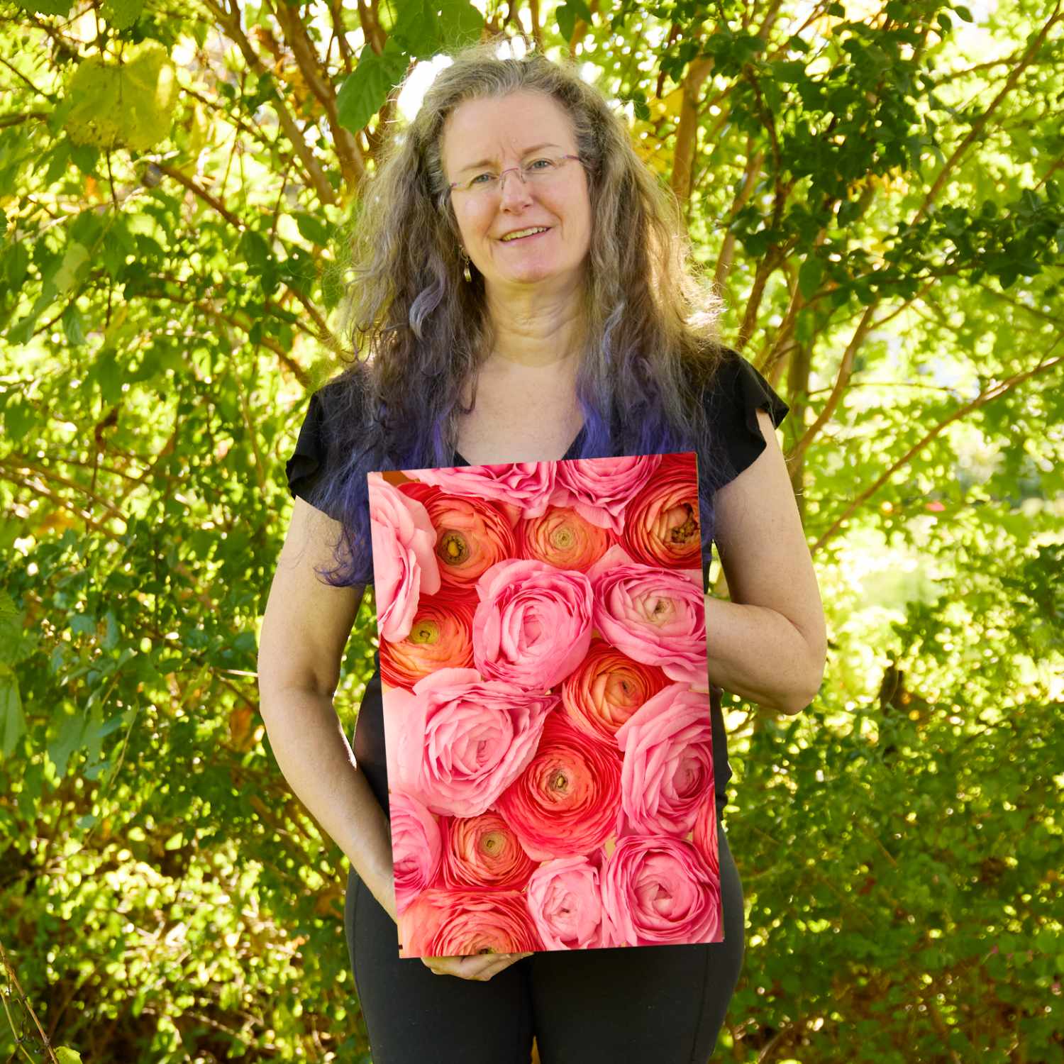 Christina Peters holding Layers of Love Ranunculus Wall Art Canvas, a vertical close-up of coral, salmon and pink ranunculus blooms packed together, showing their swirled layers of petals. She is standing outdoors in front of sunlit green trees and leafy shrubs.