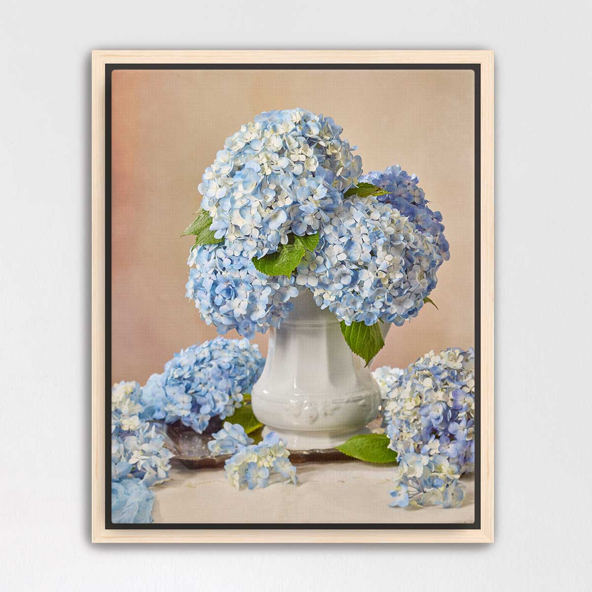 A white vase filled with blue hydrangea flowers sits on a table, surrounded by additional blue hydrangea flowers.