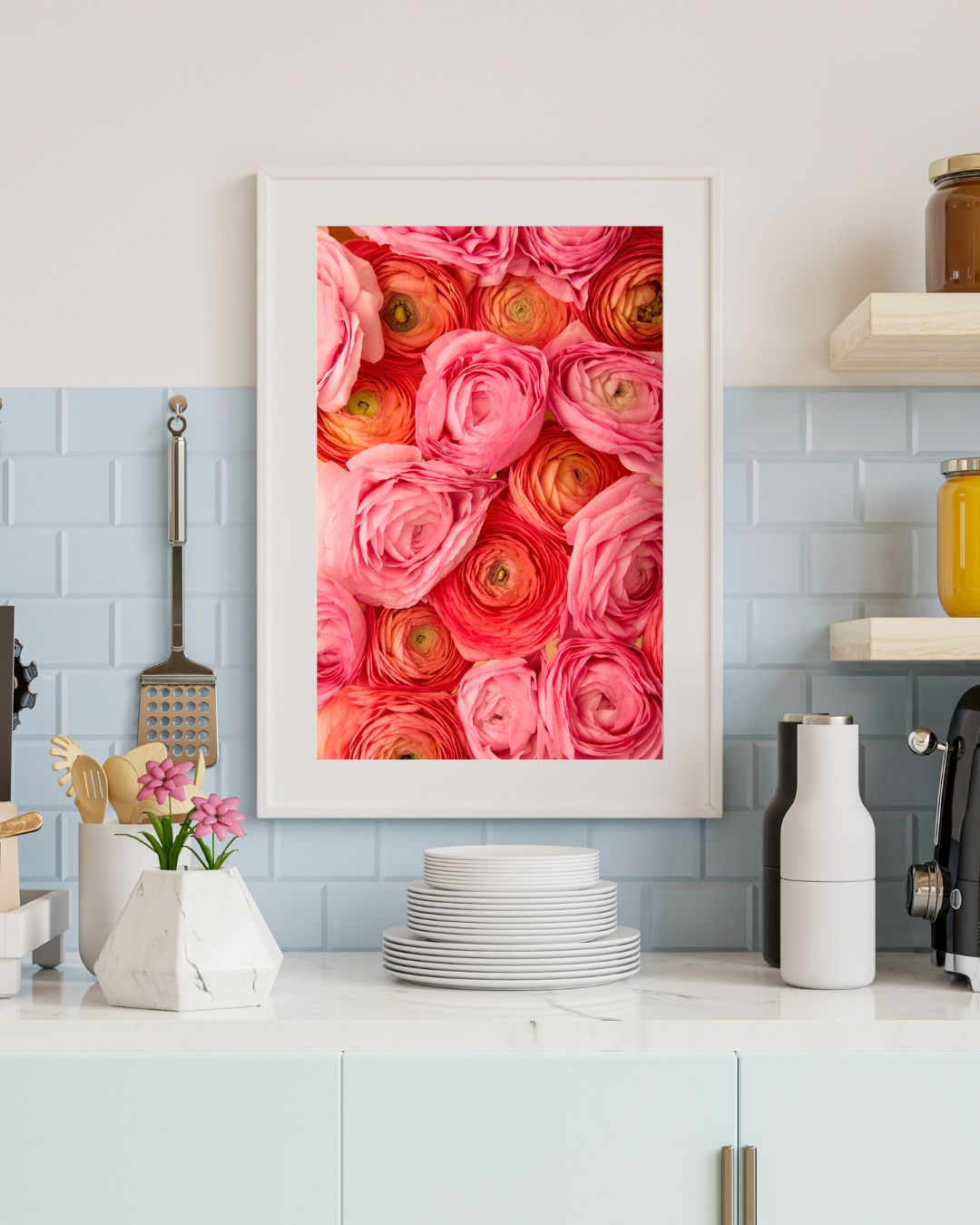 A modern kitchen with a white countertop showcases a framed Layers of Love print, depicting pink, coral and orange ranunculus from overhead. A light blue tile wall complements the counter, adorned with stacked white plates, small kitchen utensils.