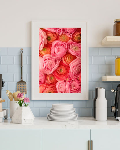 A modern kitchen with a white countertop showcases a framed Layers of Love print, depicting pink, coral and orange ranunculus from overhead. A light blue tile wall complements the counter, adorned with stacked white plates, small kitchen utensils.