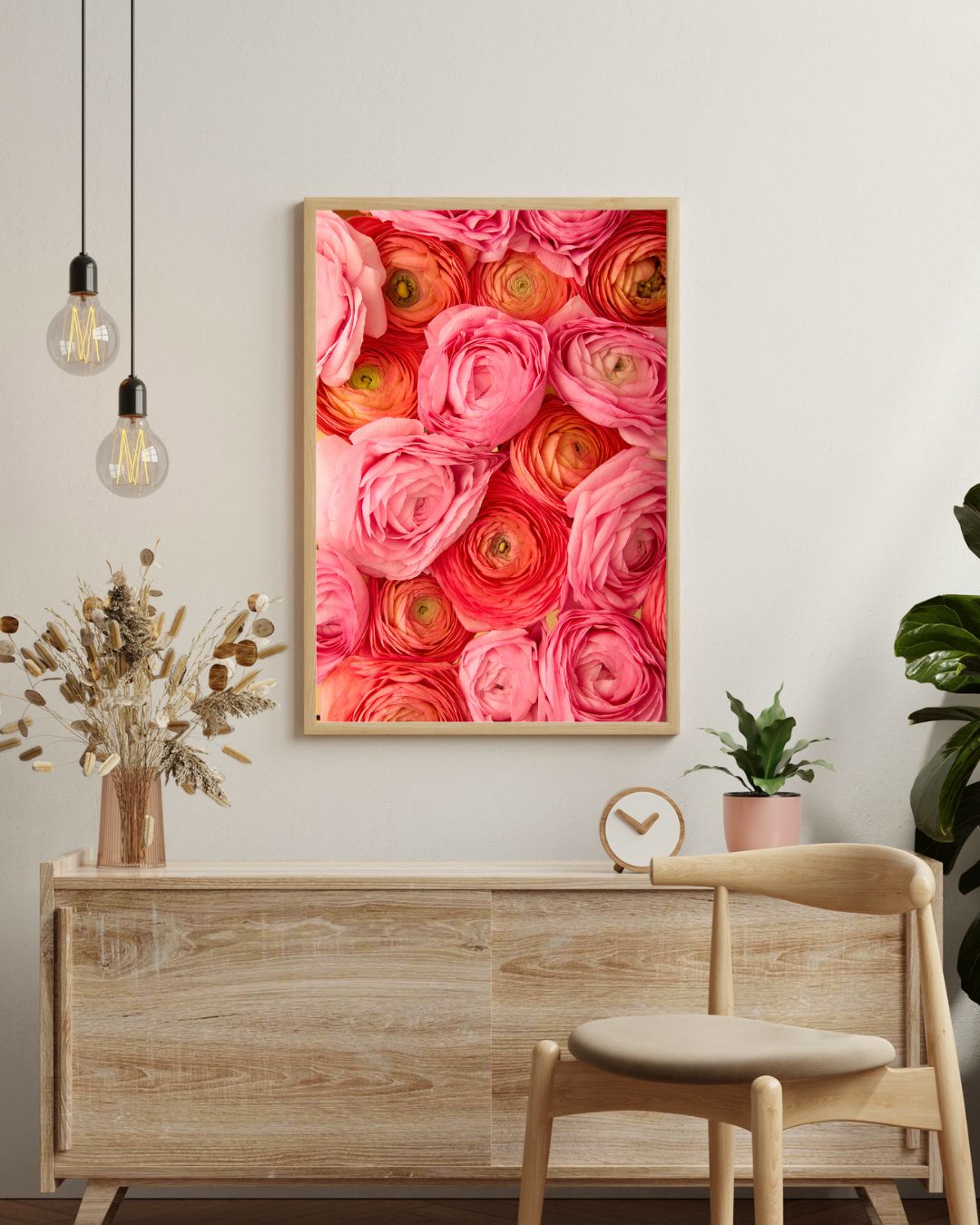 The Layers of Love canvas art in a modern thin wood frame, showcasing pink, coral and orange ranunculus, hangs above a wooden sideboard with a clock, potted plant, and dried flowers. Two pendant lights are overhead, while a wooden chair is partially visible in the foreground.