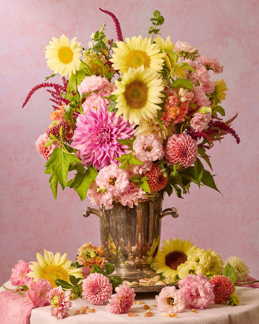 The Summers Grace bouquet features yellow sunflowers, pink dahlias, a few zinnias, pastel pink lisianthus, celosia and some greens in an antique silver champagne bucket against a soft pink background, with scattered blooms on the tablecloth. A stunning floral arrangement ideal for canvas or print wall art.