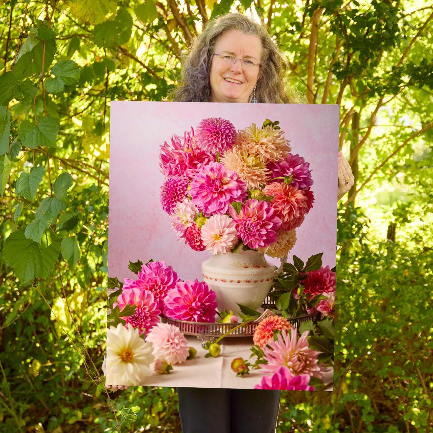 Christina Peters holding Summer Afternoon Dahlia Wall Art Canvas, a still life of hot pink, coral and peach dahlias in a white vase on a silver tray, with more dahlias around it against a soft pink backdrop. She is standing outdoors in front of sunlit green trees and leafy shrubs.
