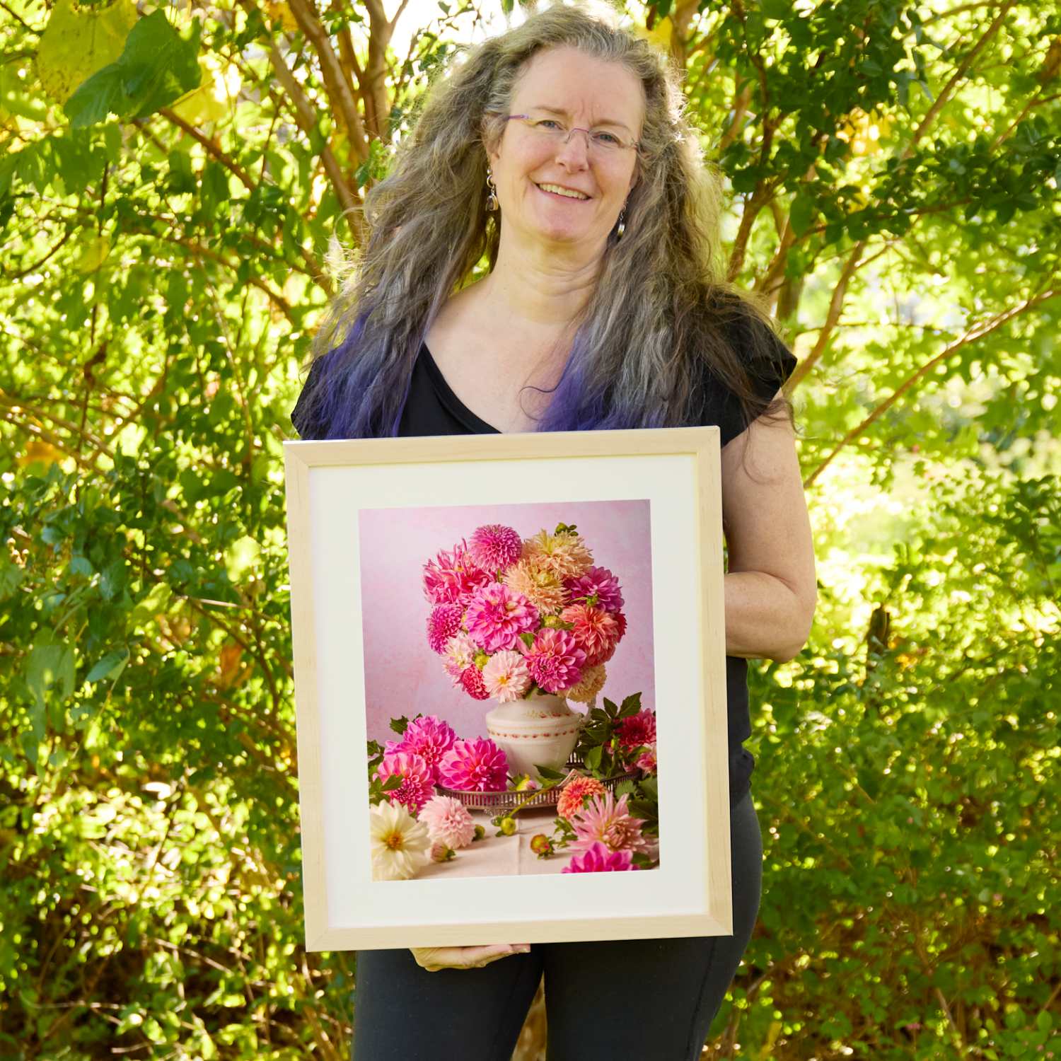 Christina Peters holding Summer Afternoon Dahlia Wall Art Print, a still life of hot pink, coral and peach dahlias in a white vase on a silver tray, with more dahlias around it against a soft pink backdrop, framed in natural wood with a white mat. She is standing outdoors in front of sunlit green trees and leafy shrubs.