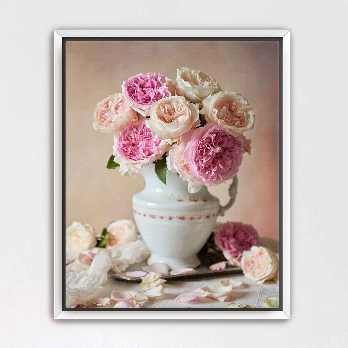 A white vase filled with pink and white roses sits on a table, surrounded by additional rose petals.