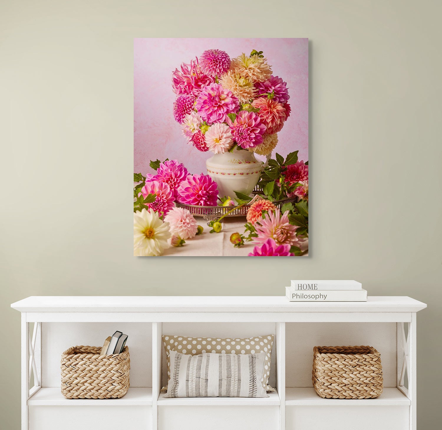 A wall with Summer Afternoon canvas depicting a vintage porcelain pitcher filled with pink, orange, and cream dahlias. Below, a white console table holds three woven baskets and two book stacks labeled Home and Philosophy.