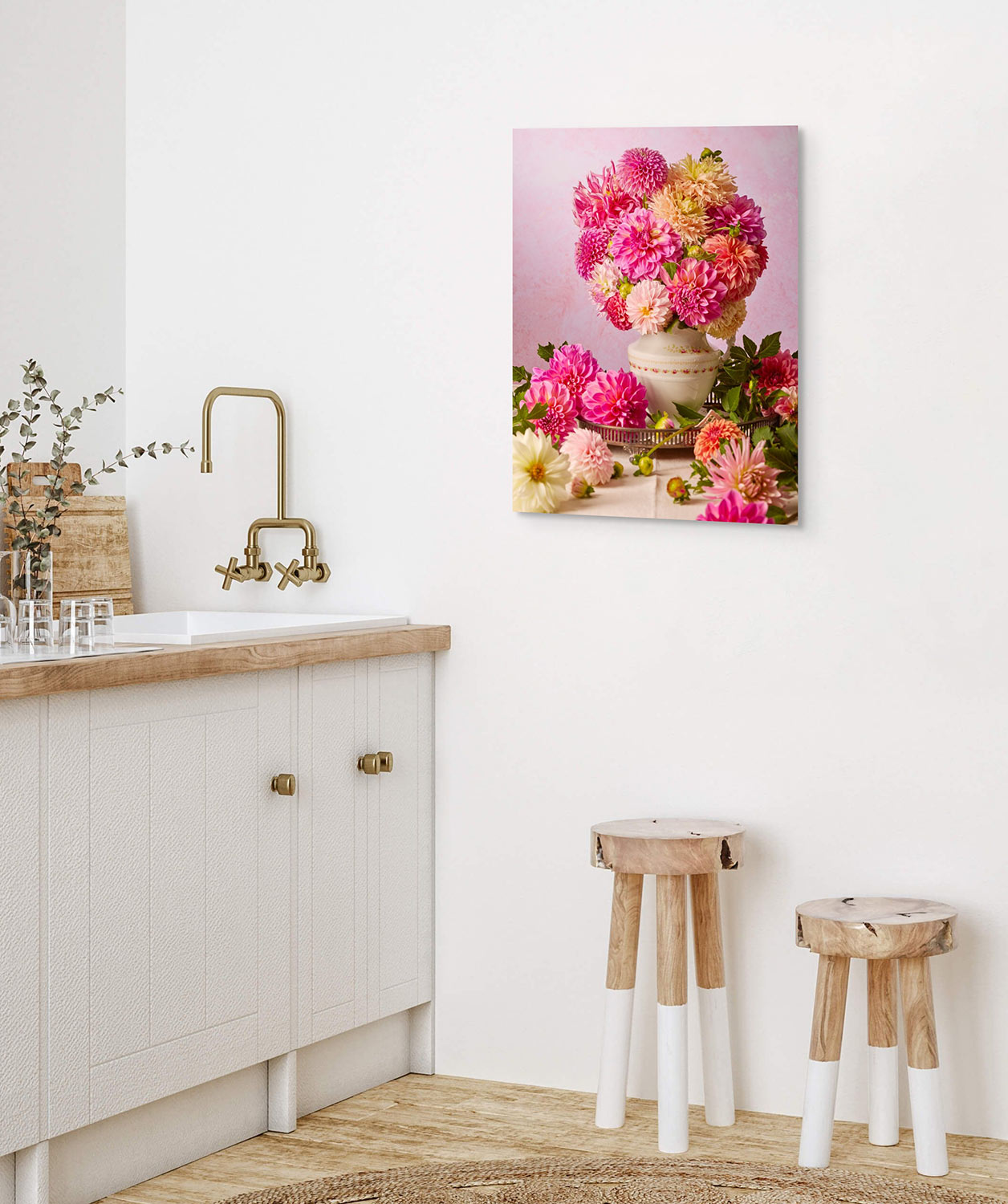 A minimalist kitchen with light wood and white tones showcases a sink with gold fixtures and two wooden stools. Wall art named Summer Afternoon displays vibrant pink flowers in a vintage porcelain pitcher, complemented by an olive branch in a vase on the counter.