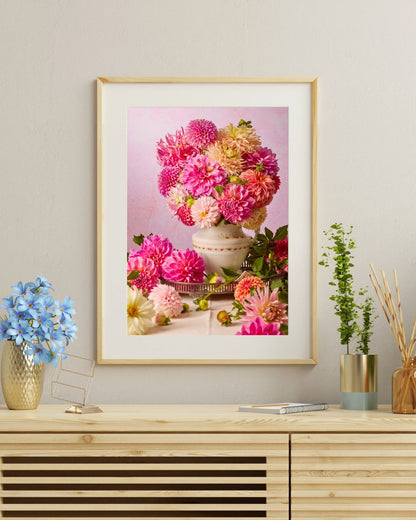 A framed Summer Afternoon photo of pink, cream and orange dahlias hangs on a beige wall above a light wood shelf. The shelf also holds a vintage porcelain pitcher with blue flowers, a silver pot with a small green plant, and a book.