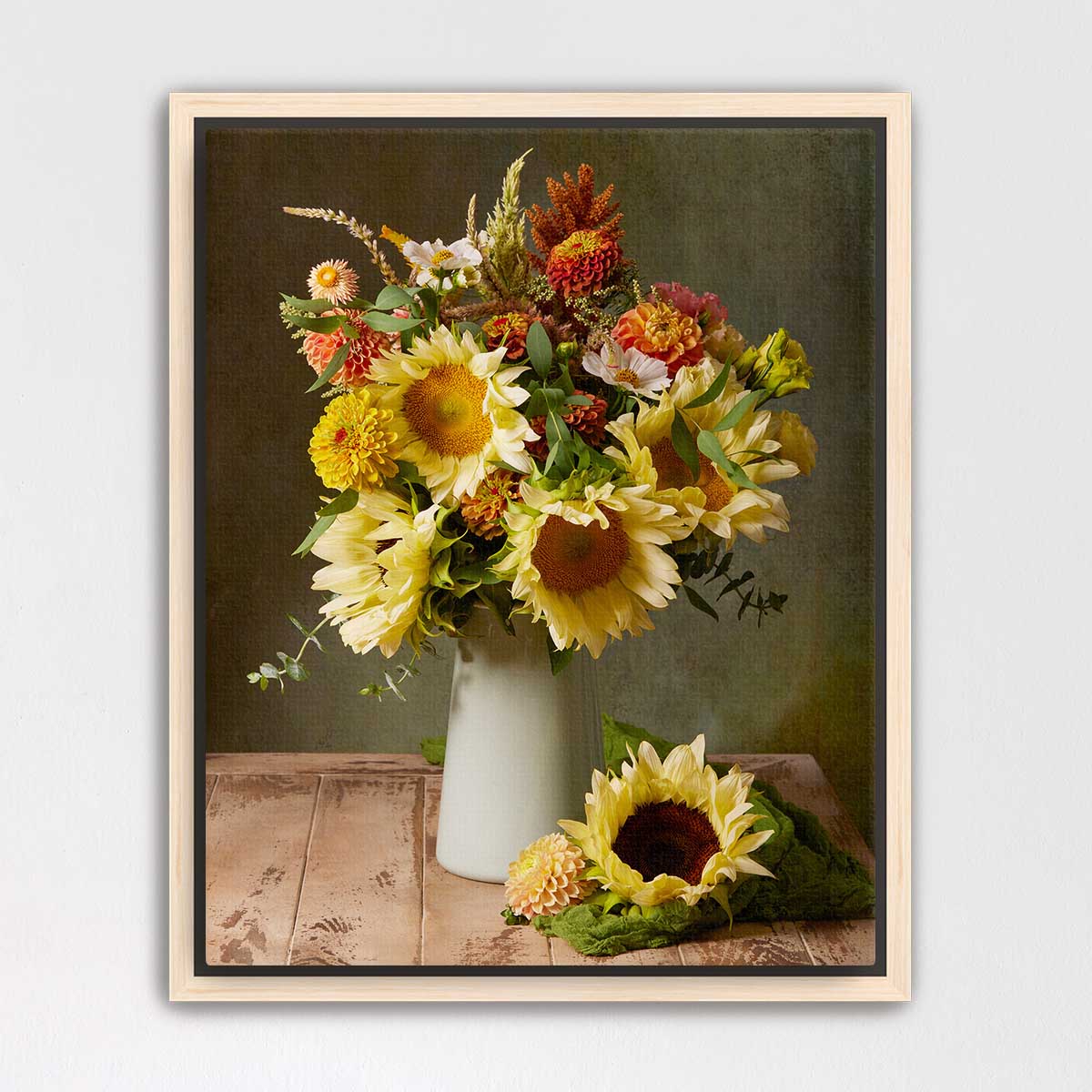 A framed still life photograph depicting a bouquet of vibrant yellow sunflowers in a white vase, placed on a wooden surface.