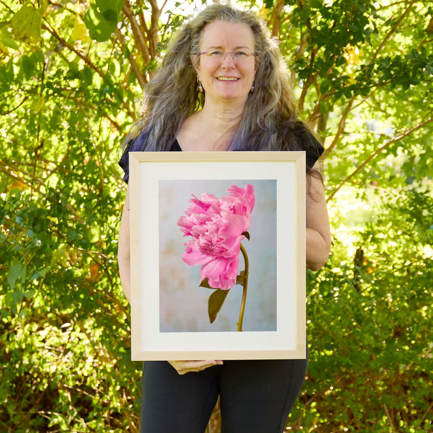 Christina Peters holding Miss Bashful Peony Wall Art Print, a single pink peony in profile, its ruffled bloom nodding on a curved stem against a soft pale blue textured backdrop, framed in natural wood with a white mat. She is standing outdoors in front of sunlit green trees and leafy shrubs.