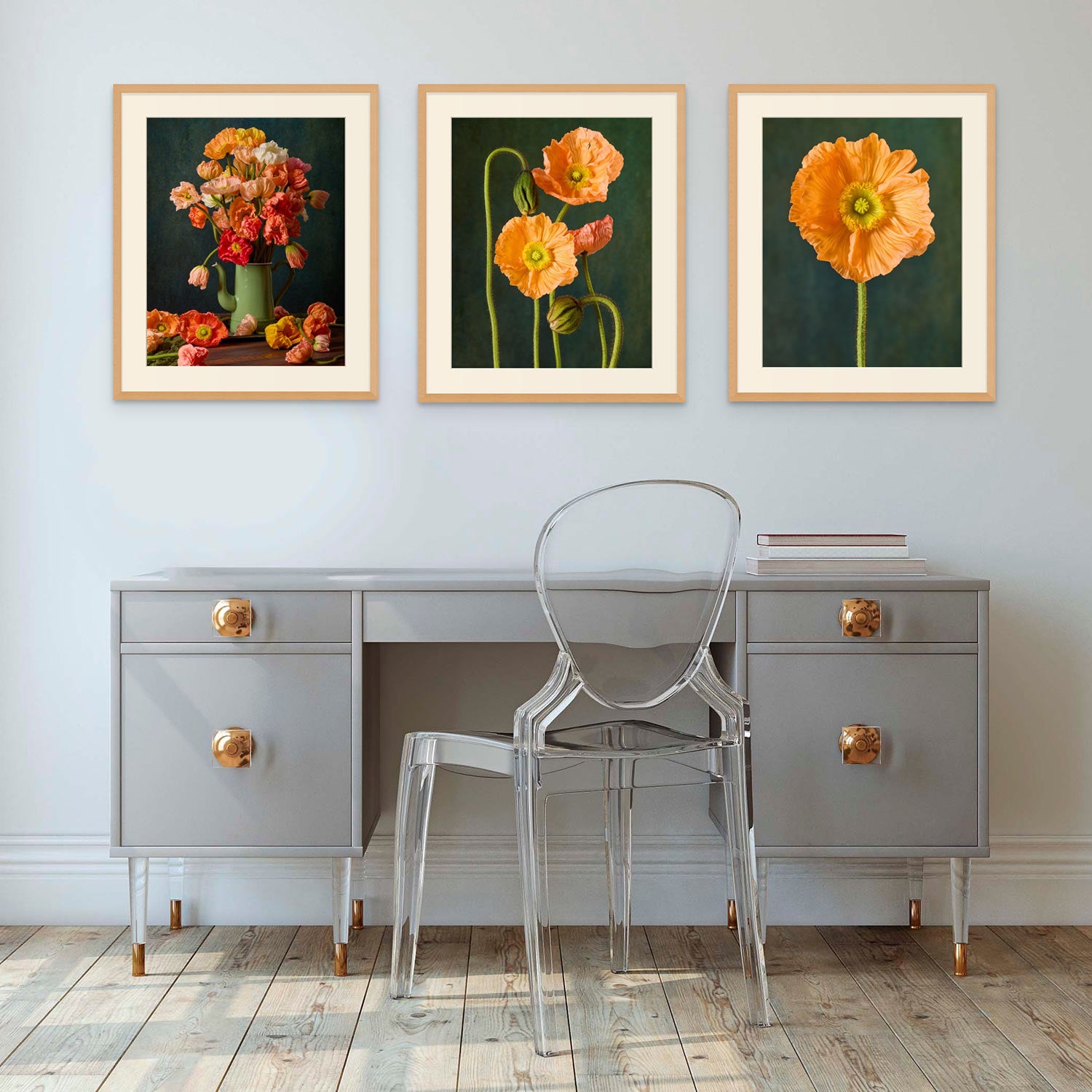 A modern room with a gray desk adorned in gold accents and a transparent chair showcases the Poppy Sisters Portraits - 3 Piece art set featuring Italian pastel poppies above it, all enhanced by light wood flooring to create an elegant chic space.