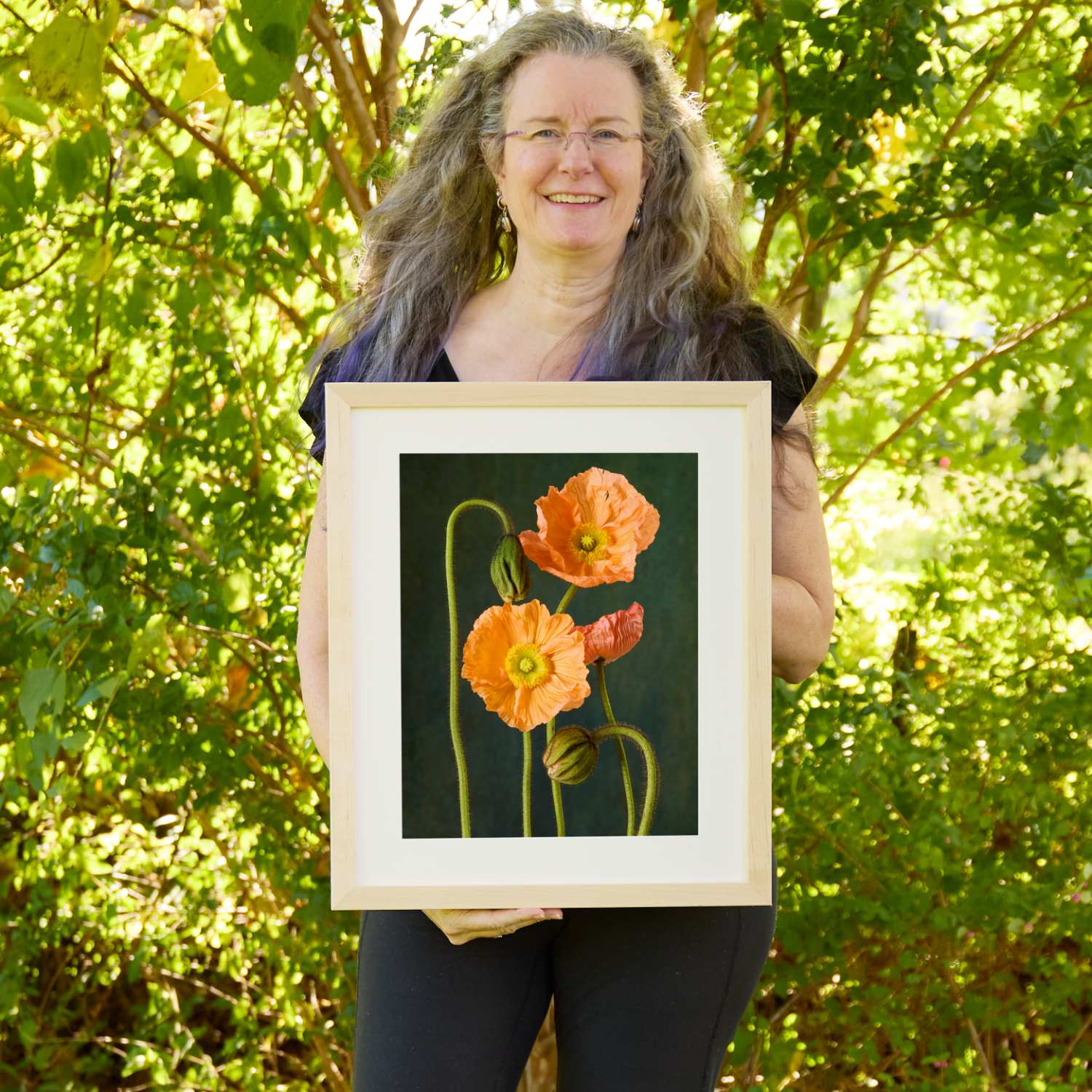 Christina Peters holding The Poppy Sisters Wall Art Print, three peach and coral Iceland poppies with fuzzy curving stems and closed buds against a deep green backdrop, framed in natural wood with a white mat. She is standing outdoors in front of sunlit green trees and leafy shrubs.