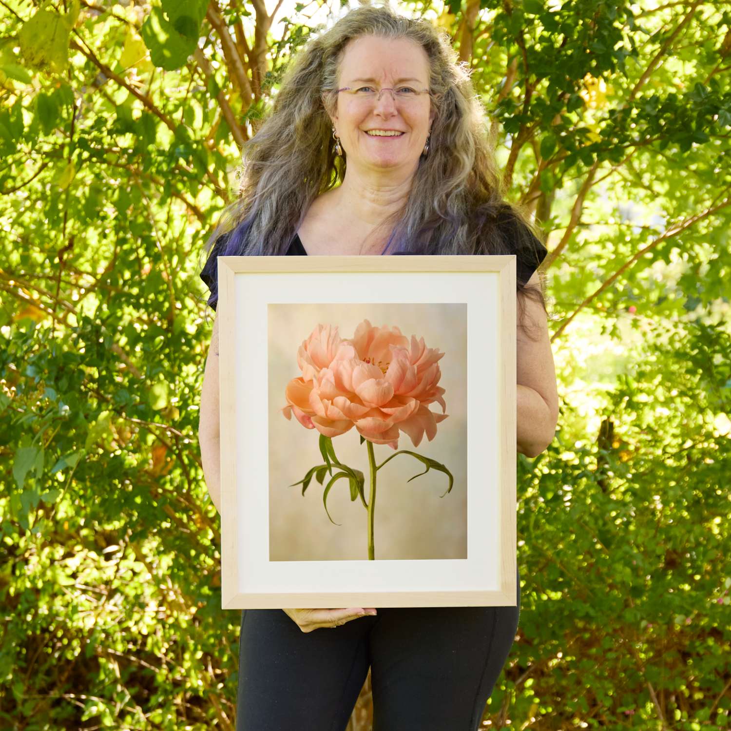 Christina Peters holding Coral Charm Peony Portrait 3 Wall Art Print, a single soft peach peony in full bloom on a stem with arching leaves against a soft cream backdrop, framed in natural wood with a white mat. She is standing outdoors in front of sunlit green trees and leafy shrubs.