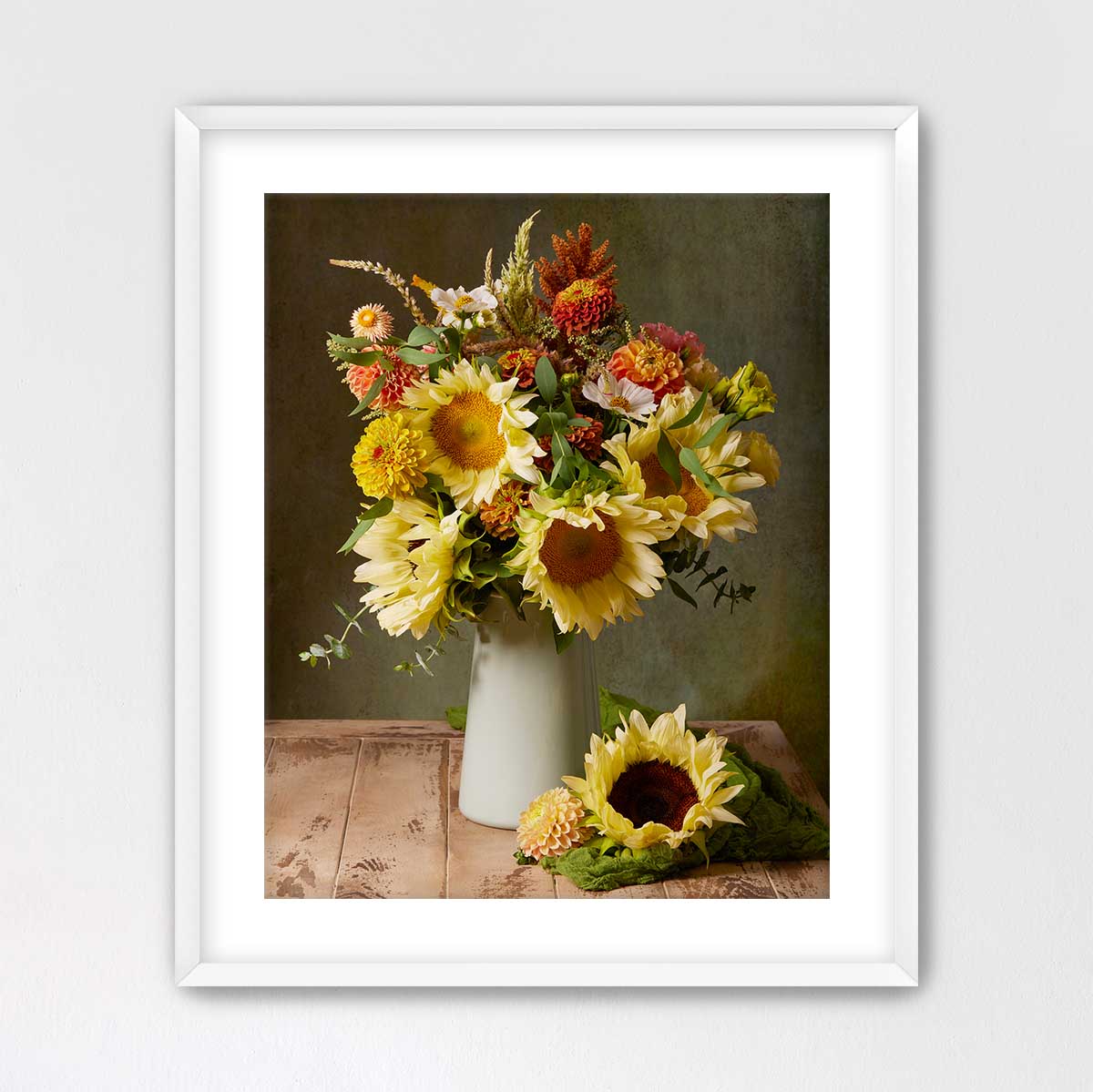 A framed image of a bouquet of yellow sunflowers and other flowers in a white vase, placed on a wooden surface.