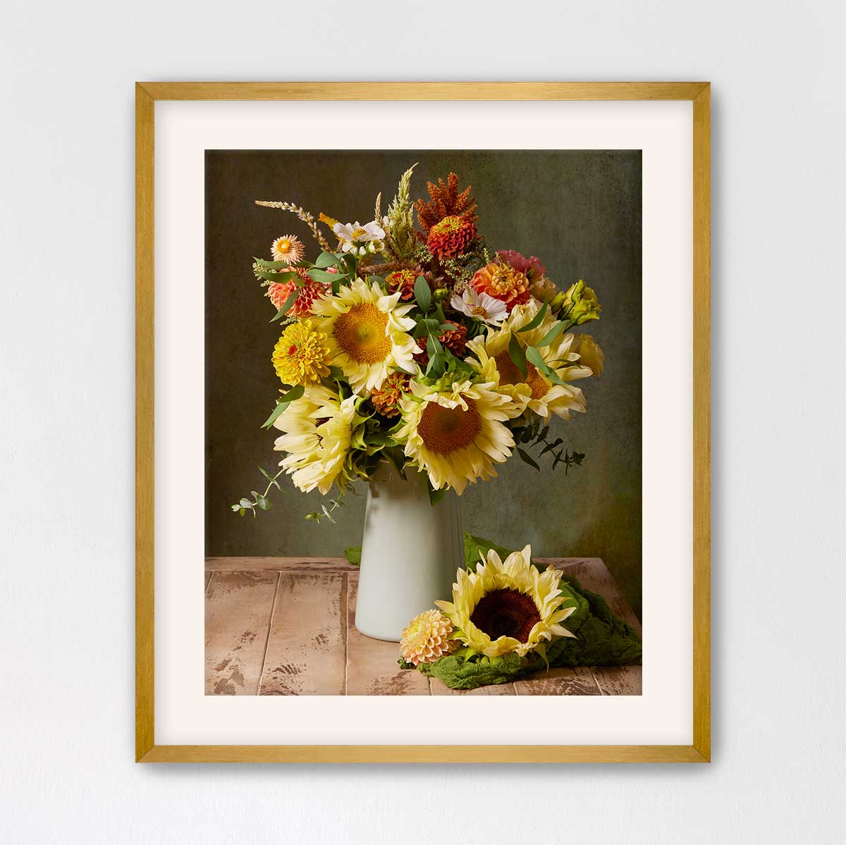 A framed photograph of a bouquet of yellow sunflowers and other flowers in a white vase, placed on a wooden surface.