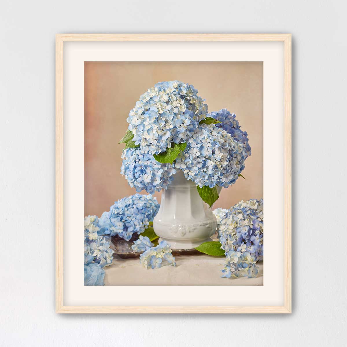 A framed image of a white vase filled with blue hydrangea flowers, surrounded by additional blue hydrangea flowers on a table.