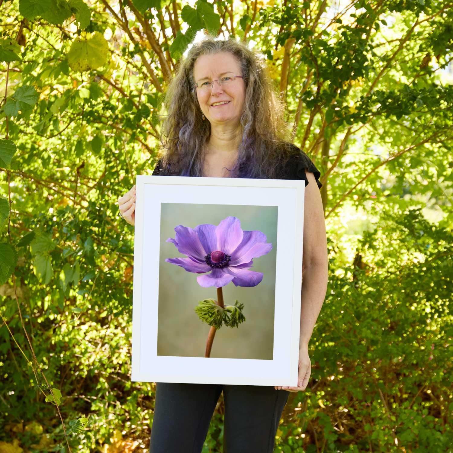 Christina Peters holding Her Highness Anemone Wall Art Print, a single lavender anemone with a dark center on a tall stem with a ruff of green leaves against a soft sage backdrop, framed in white with a white mat. She is standing outdoors in front of sunlit green trees and leafy shrubs.