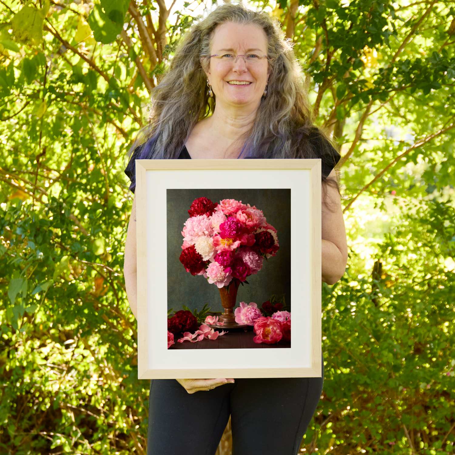 Christina Peters holding Passion In Bloom Peony Wall Art Print, a still life of crimson, magenta, pink and white peonies in a copper vase, with loose blooms and petals on the table against a dark backdrop, framed in natural wood with a white mat. She is standing outdoors in front of sunlit green trees and leafy shrubs.