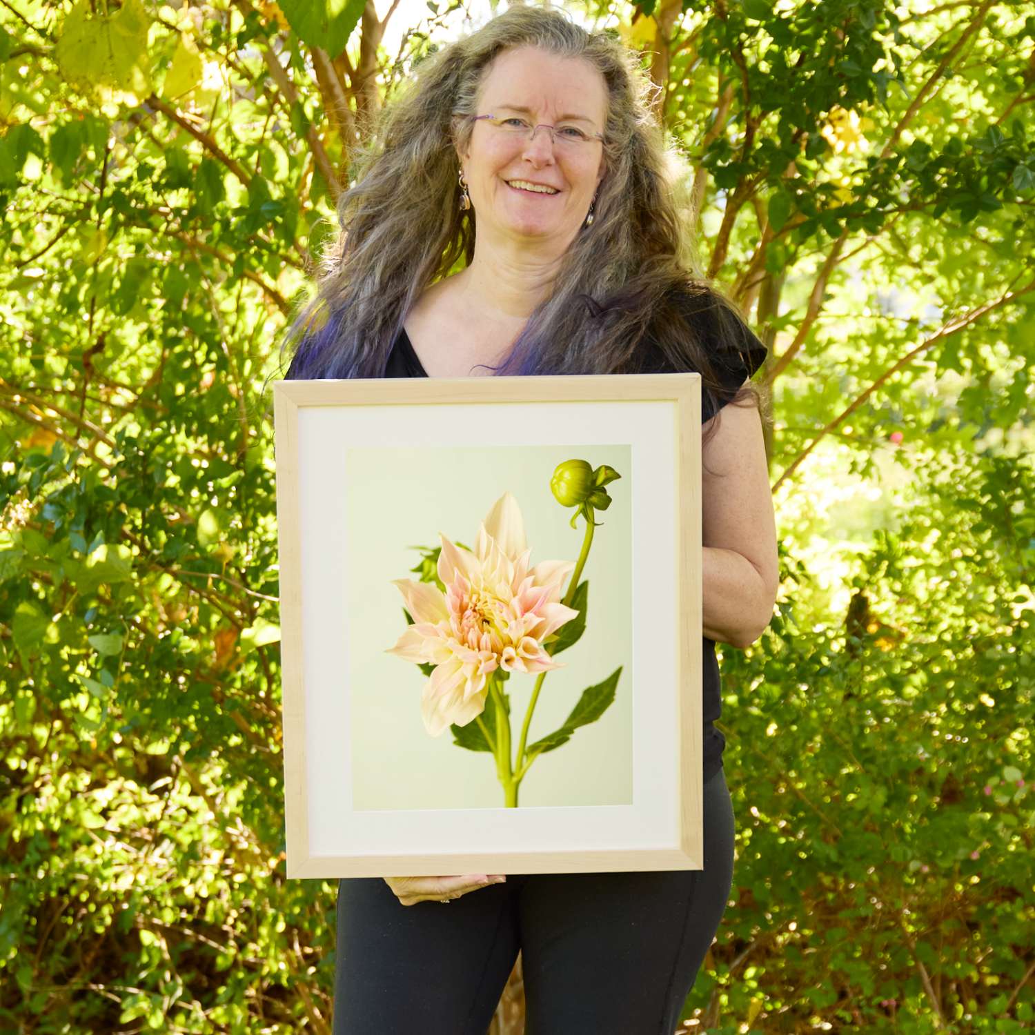 Christina Peters holding Peaches and Cream Dahlia Wall Art Print, a single blush and cream dinnerplate dahlia with a green bud on a branching stem against a soft pale green backdrop, framed in natural wood with a white mat. She is standing outdoors in front of sunlit green trees and leafy shrubs.