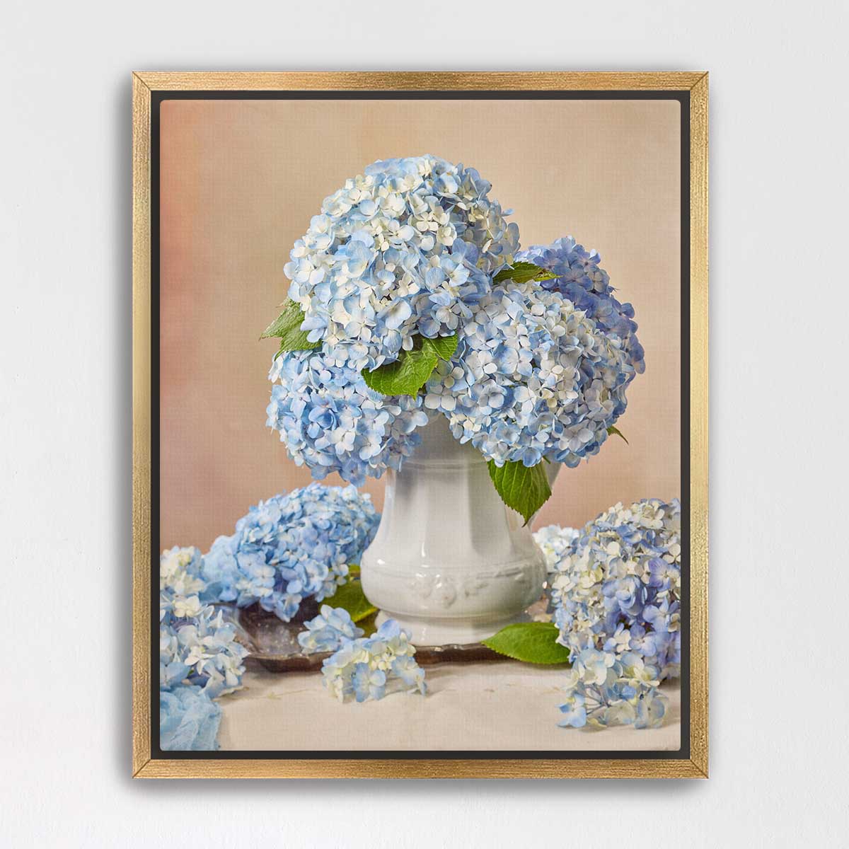 A white vase filled with blue hydrangea flowers sits on a table, surrounded by additional blue hydrangea flowers.