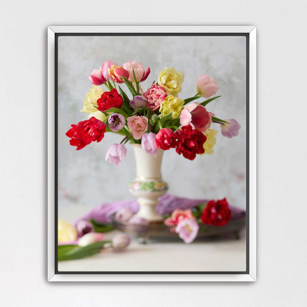 A white vase filled with a vibrant bouquet of red, pink, yellow, and white flowers sits on a table, surrounded by additional flowers and a purple cloth.