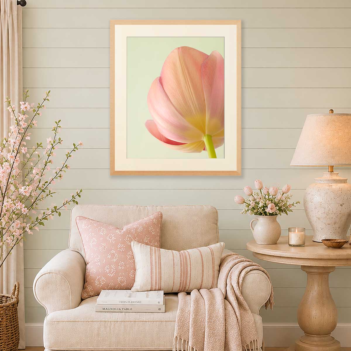 A framed art print of a soft pink tulip with a pale green background, displayed on a light green paneled wall above a beige armchair in a styled living room setting.
