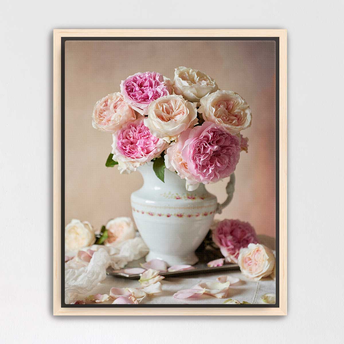 A white vase filled with pink and white roses sits on a table, surrounded by additional rose petals.