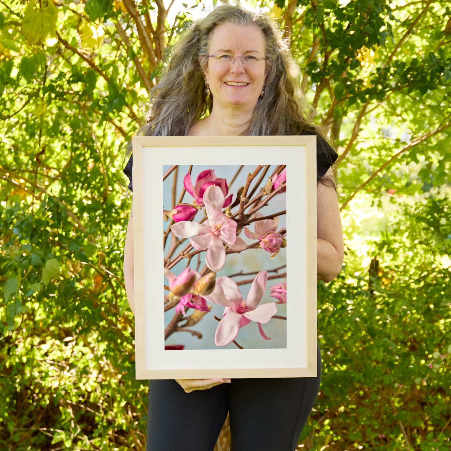 Christina Peters holding Magnolia Duet Wall Art Print, pale pink and magenta magnolia blossoms and buds on bare branches against a soft blue sky, framed in natural wood with a white mat. She is standing outdoors in front of sunlit green trees and leafy shrubs.