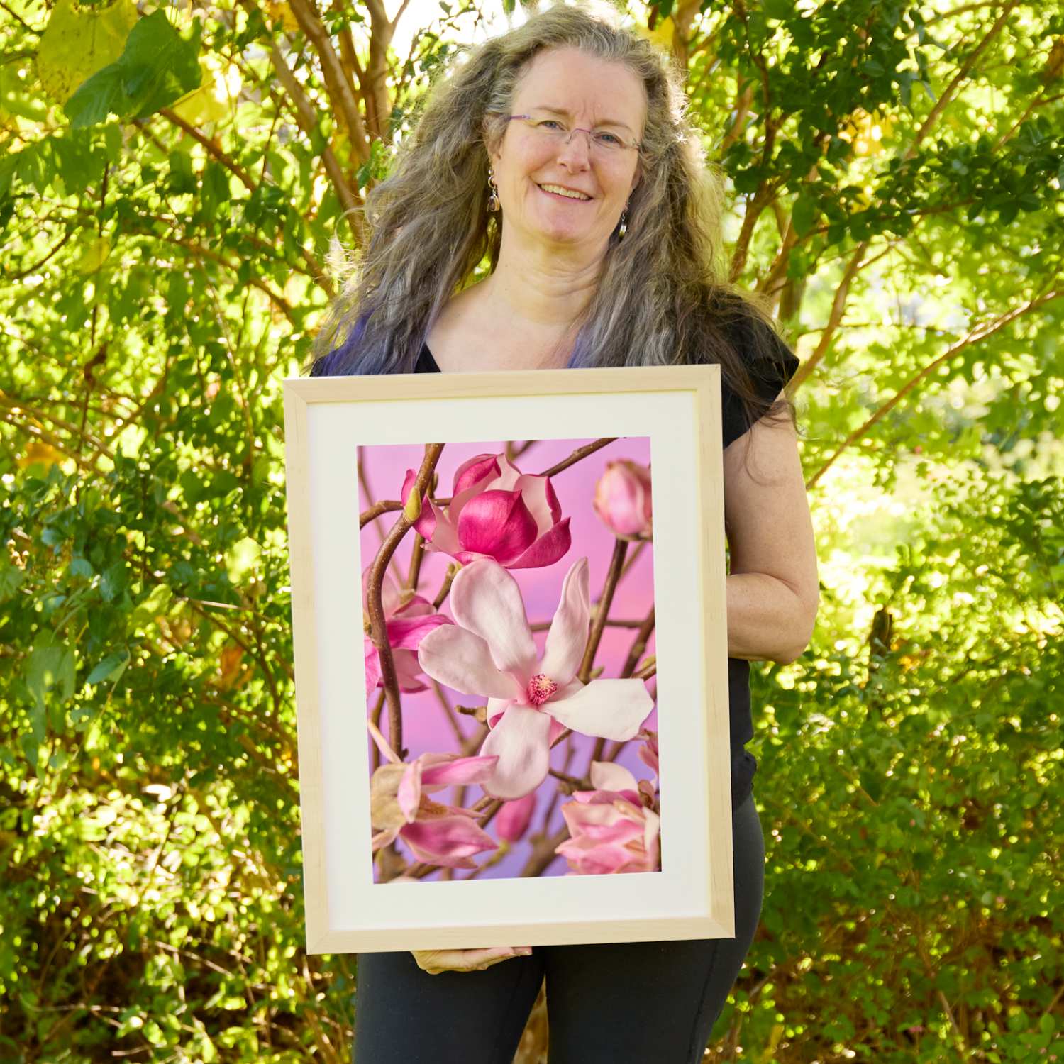 Christina Peters holding Magnolia Muse Wall Art Print, pale pink and magenta magnolia blossoms and buds on bare branches against a soft pink and lavender backdrop, framed in natural wood with a white mat. She is standing outdoors in front of sunlit green trees and leafy shrubs.