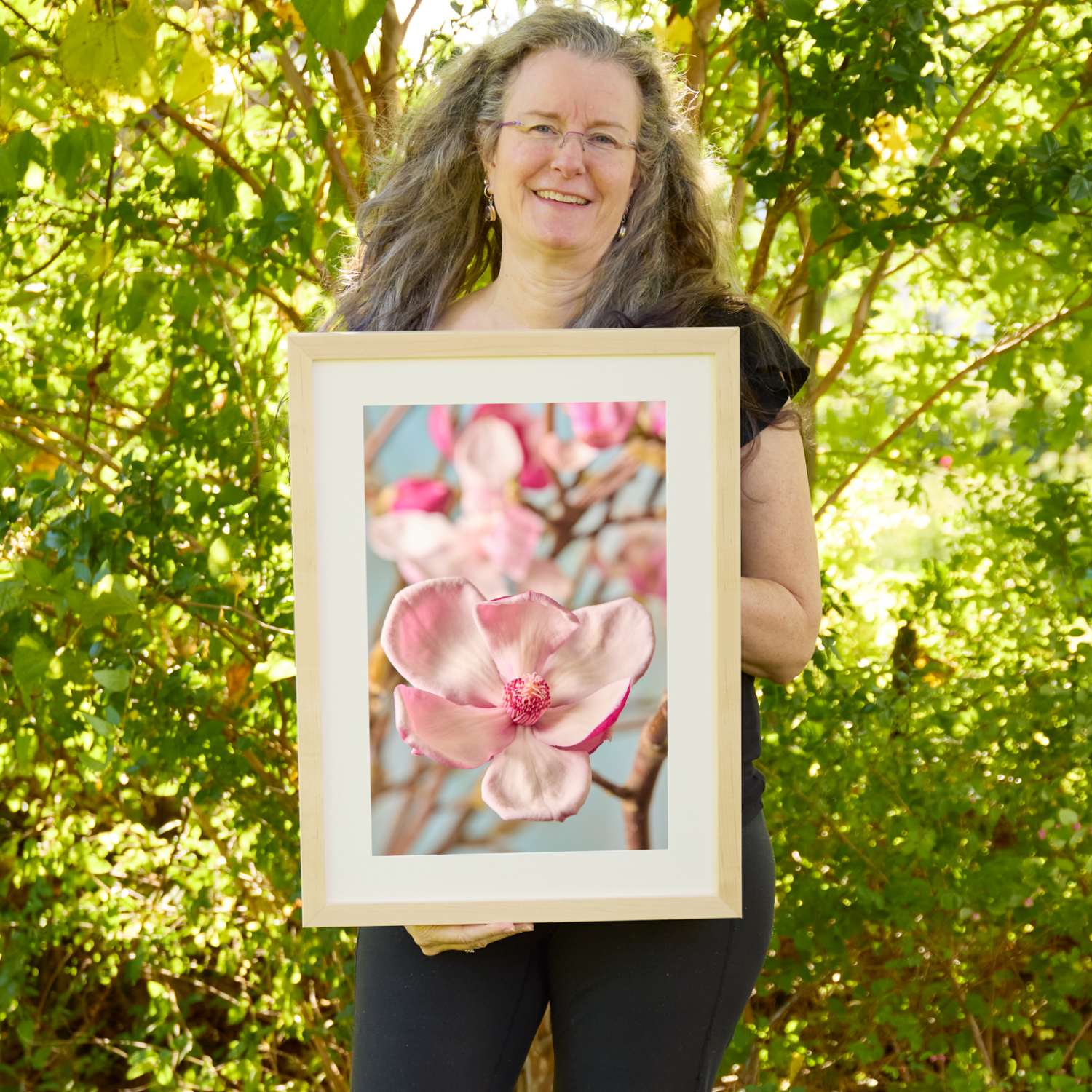 Christina Peters holding Gentle Radiance Magnolia Wall Art Print, a close-up of a single open pale pink magnolia blossom on a branch, with softly blurred blooms behind it against a pale blue sky, framed in natural wood with a white mat. She is standing outdoors in front of sunlit green trees and leafy shrubs.