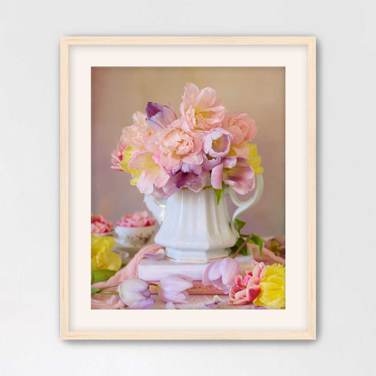 A white vase filled with a variety of pink and yellow flowers sits on a pink surface, surrounded by additional flowers.