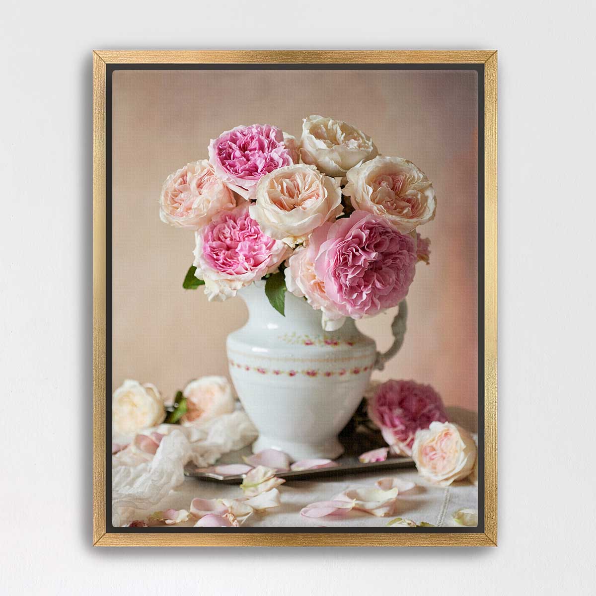 A white vase filled with pink and white roses sits on a table, surrounded by additional rose petals.