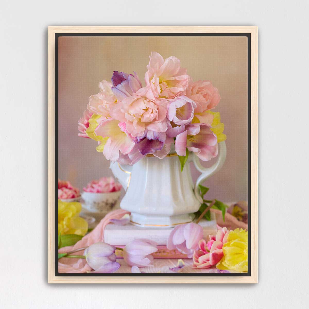 A white vase filled with a bouquet of pink and yellow flowers sits on a table, surrounded by additional flowers and a book.