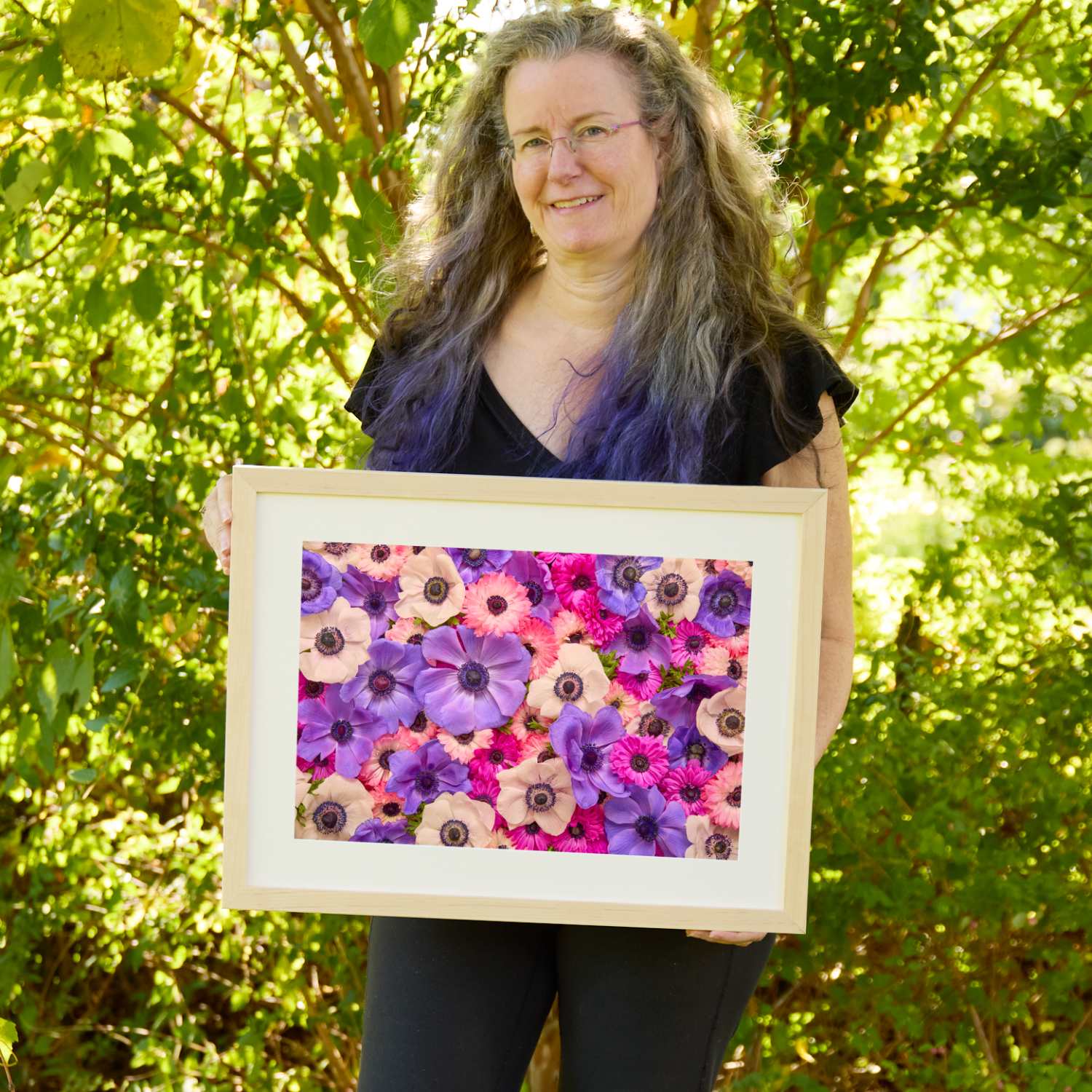 Christina Peters holding Purple Passion Horizontal Anemone Wall Art Print, a horizontal close-up of purple, magenta, pink and white anemones packed together, framed in natural wood with a white mat. She is standing outdoors in front of sunlit green trees and leafy shrubs.